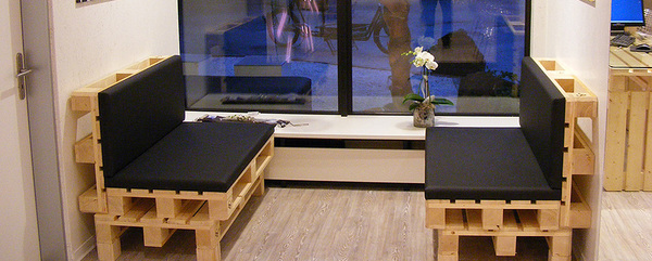 Sitzbank aus Paletten mit schwarzen Polstern steht in modernem, hellen Wartebereich.